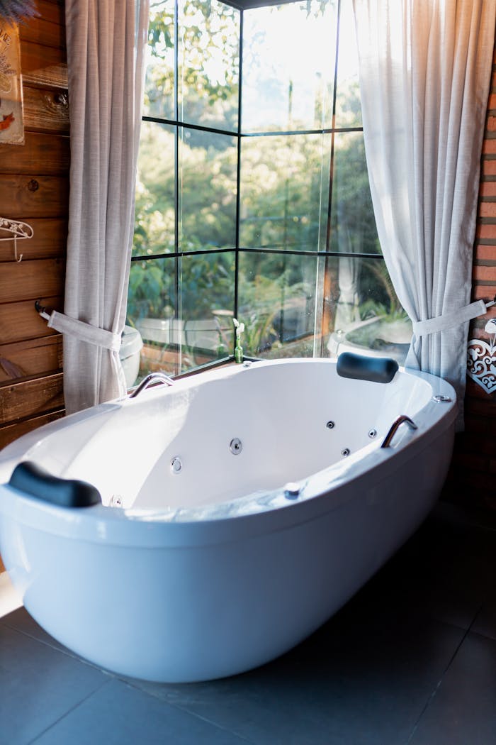 about-01 Spacious white bathtub in a sunlit bathroom with large windows and curtains, offering a serene escape.